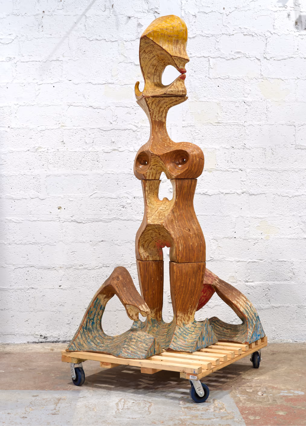 Tall abstract wooden sculpture resembling a stylized human figure with exaggerated features, including a prominent head, torso with carved breast shapes, and elongated legs, standing on a wooden platform with wheels against a white brick wall.