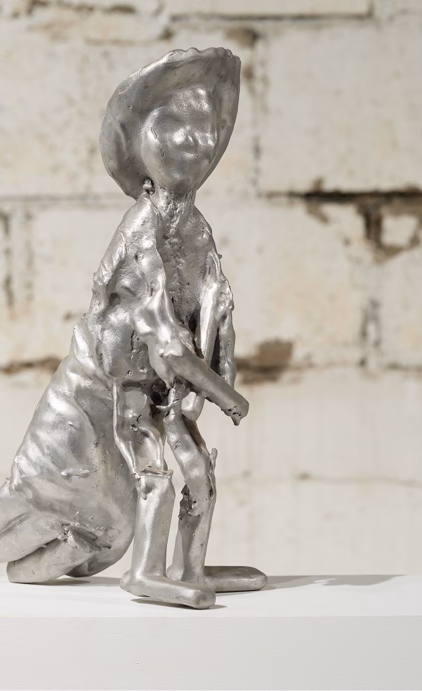 Shiny silver metallic abstract sculpture resembling a person with a large headpiece and elongated limbs seated on a white surface against a light brick wall background.