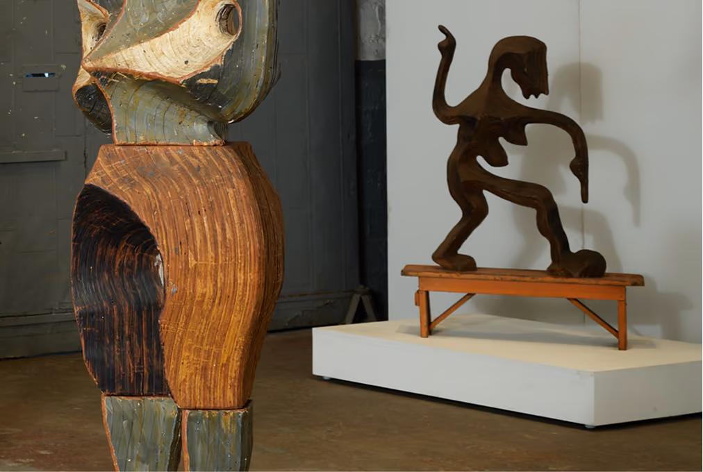 Two wooden sculptures in an indoor gallery space; the left sculpture features abstract, curved shapes with textured wood grain in brown, black, and green tones, while the right sculpture depicts a dark brown figure with exaggerated limbs on a small wooden bench, casting a shadow on a white platform.