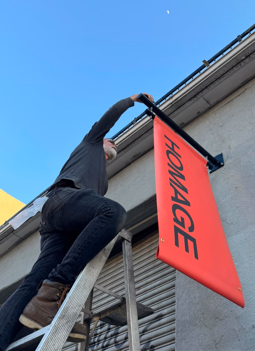 Man wearing dark clothes and brown boots climbing a metal ladder to hang or adjust a red vertical banner with the word 'HOMAGE' in black letters on the side of a gray building against a clear blue sky.
