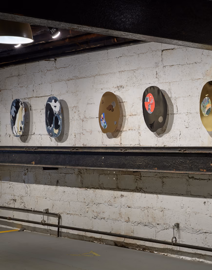 A series of abstract mixed-media circular artworks mounted on a white brick wall in an industrial-like space with metal beams and pipes below.