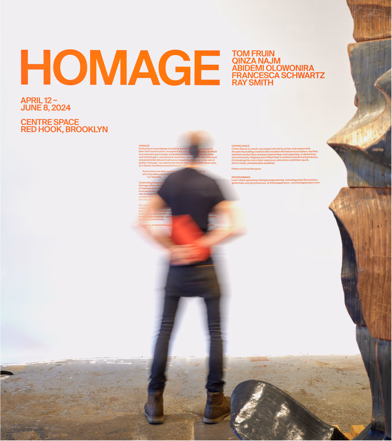 Blurred figure of a person standing with hands behind their back looking at a white wall with orange text about an art exhibition titled HOMAGE, held April 12 to June 8, 2024, at Centre Space in Red Hook, Brooklyn, with artist names on the right side and a large wooden sculpture partially visible on the right.