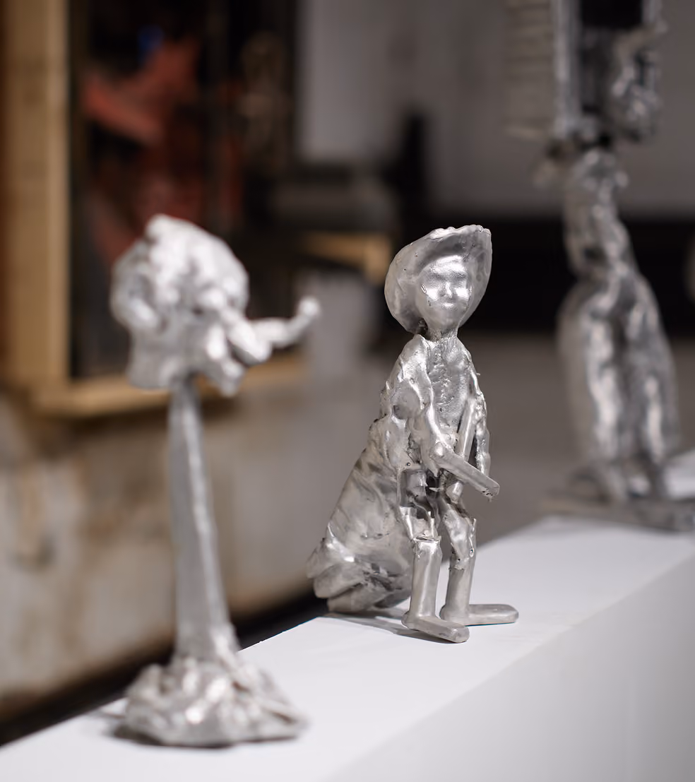 Three small, abstract silver metal sculptures arranged on a white surface with a blurred background, featuring a figure with a cloak and helmet in the center.