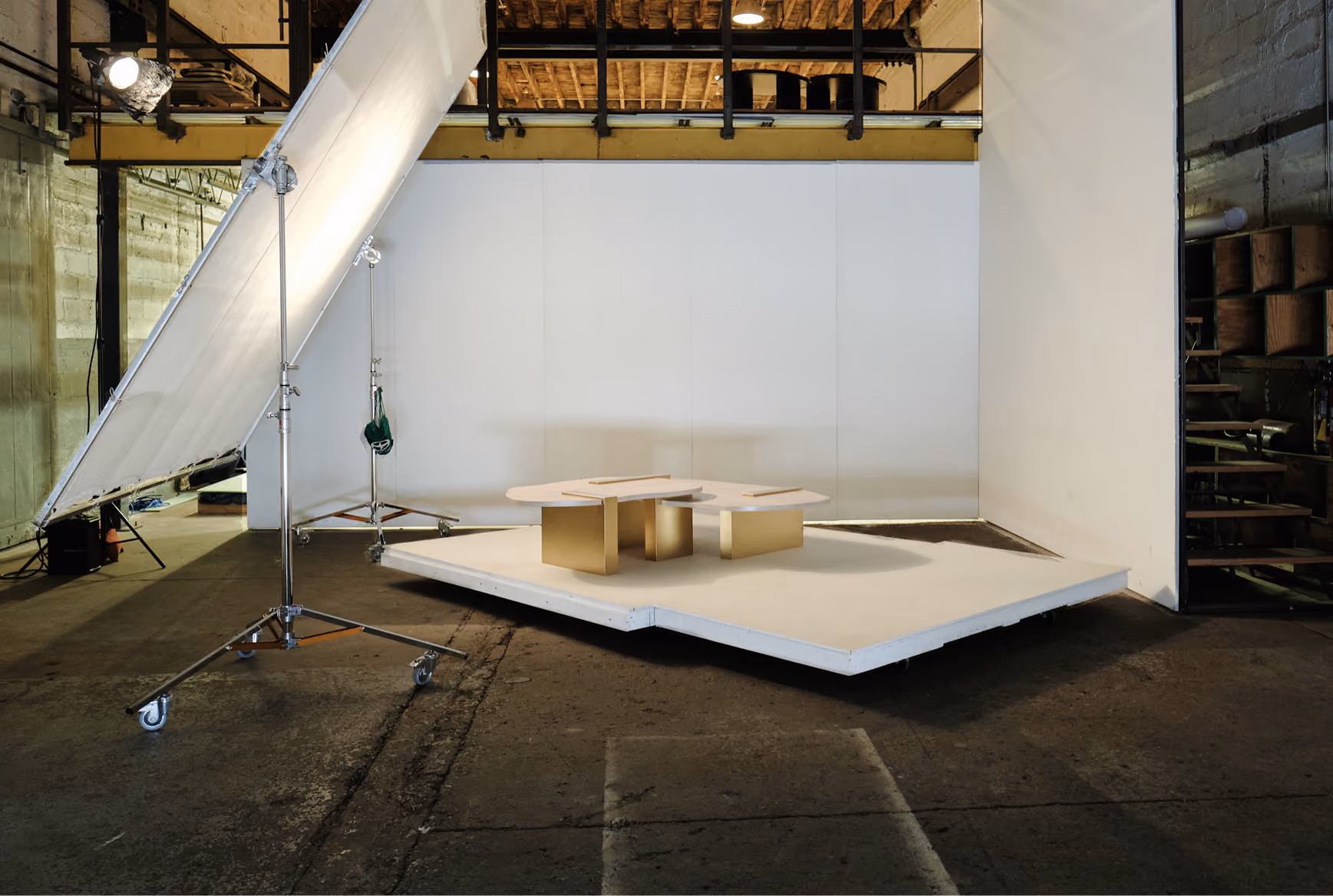 Photography studio setup with a large angled white reflector on wheels illuminating a set of modern geometric tables with gold and cream bases on a white platform against a plain white backdrop in an industrial space.