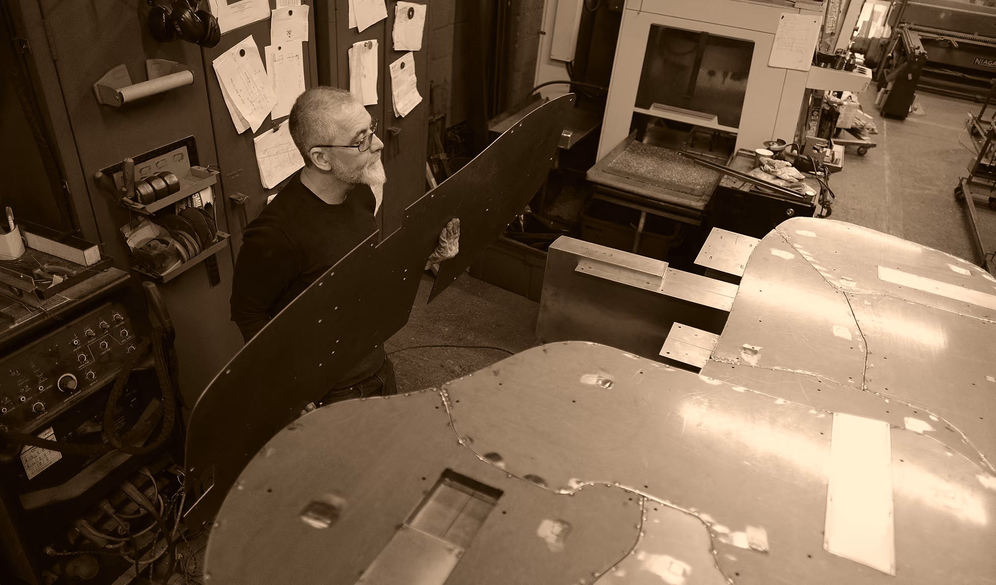 Man with glasses and a beard working in a workshop, holding a large black metal piece near a metallic aircraft wing structure, surrounded by workbenches and tools.
