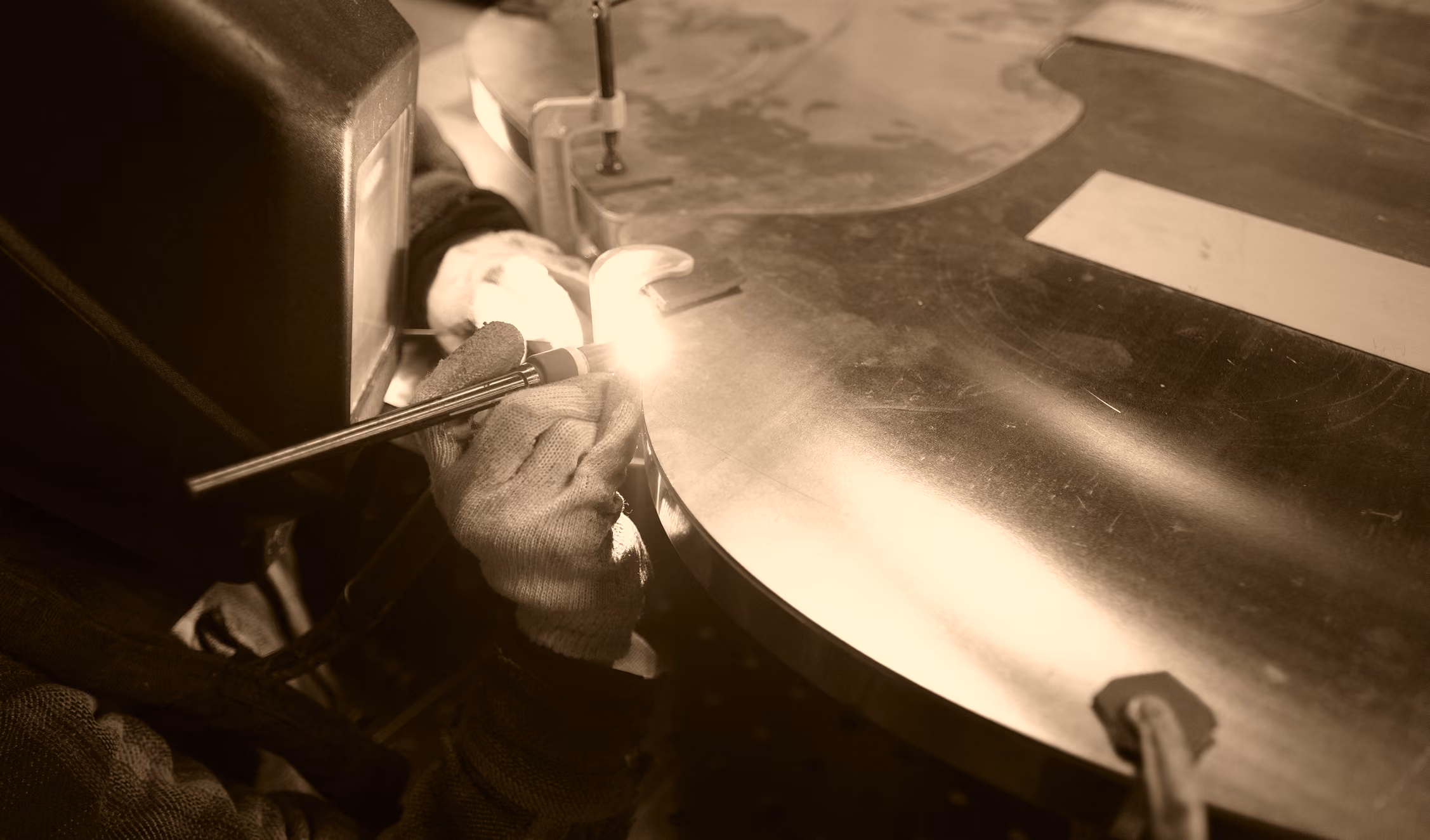 Person wearing gloves welding or cutting a large curved metal sheet with a bright spark from the welding torch.
