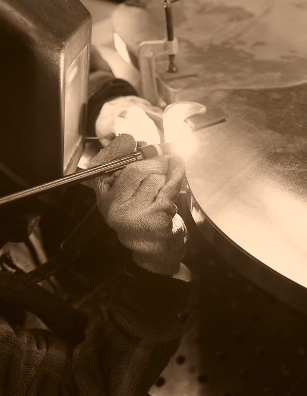Person welding with a torch on a round metal surface, wearing protective gloves and a welding helmet.