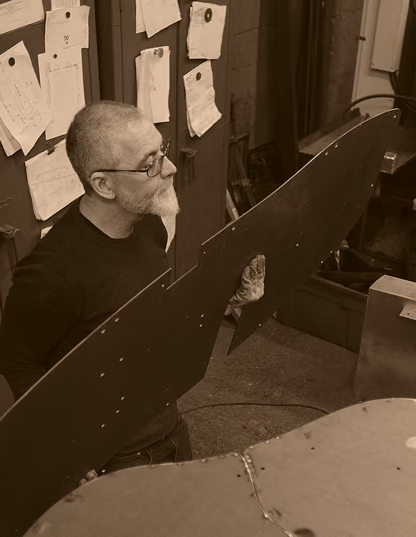 Man with glasses and beard holding a large metal sheet in a workshop with papers pinned on the wall behind him.