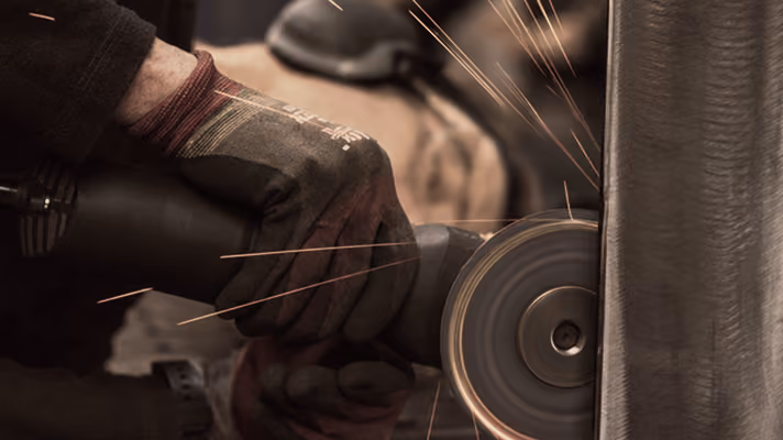 Close-up of a gloved hand holding an angle grinder producing sparks while sharpening or grinding a metal surface.
