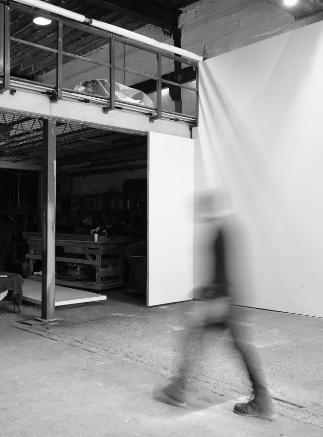 Black and white photo of a blurred figure walking across an industrial-style interior with concrete floors, a railing above, and a partially open door revealing a workshop with tools and furniture inside.