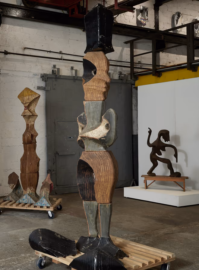 Three abstract wooden sculptures indoors on wooden platforms with wheels; the largest in front features geometric shapes forming legs, a torso, and multiple faces stacked vertically with black, brown, and gray coloring.