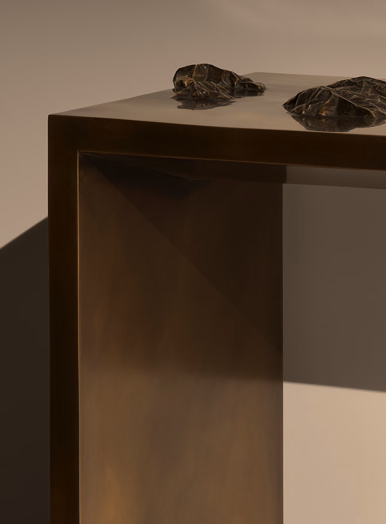 Close-up of a minimalist metallic table corner with a bronze finish and two textured rock-like decorative objects on its surface, casting soft shadows.