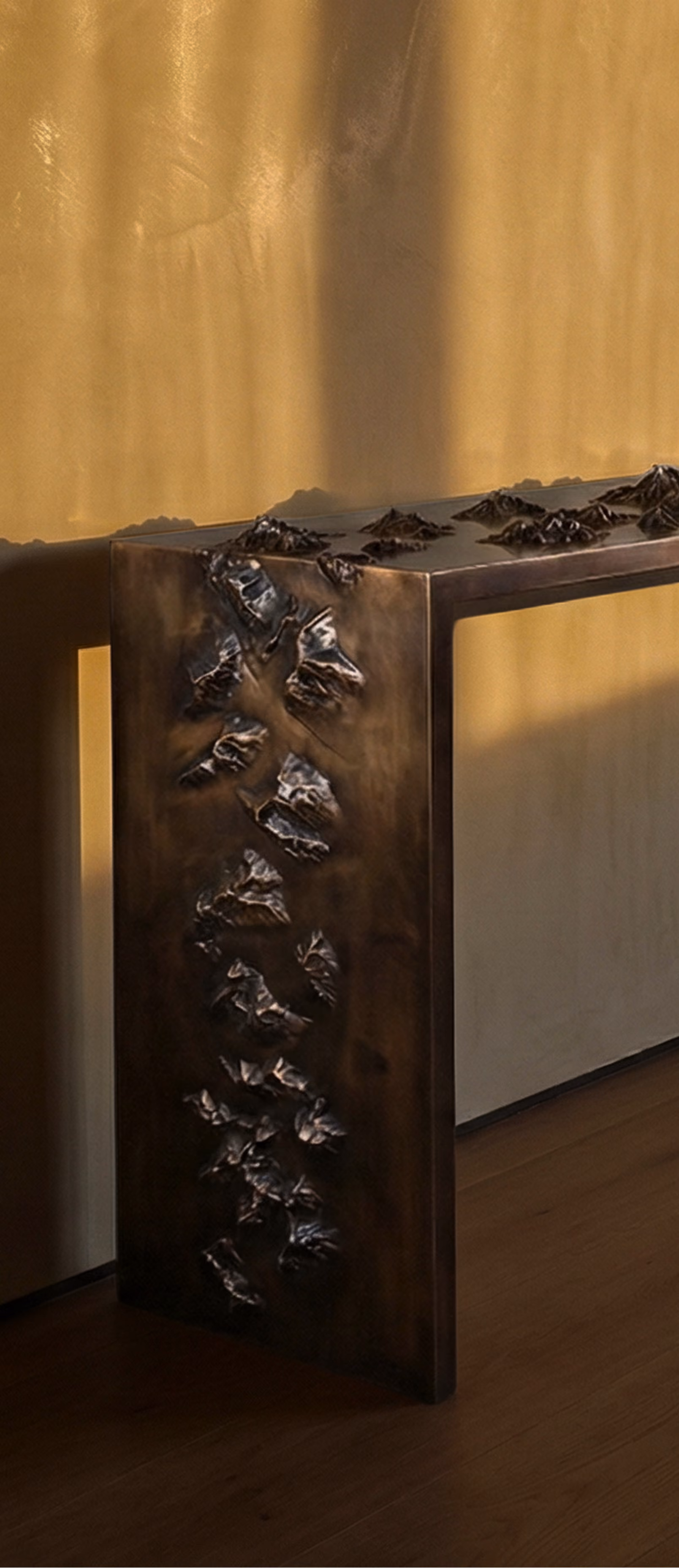 Bronze or dark metal sculpture shaped like an angular table with textured, rugged mountain-like formations on the vertical and horizontal surfaces, illuminated by warm light casting shadows on a plain wall and wooden floor.
