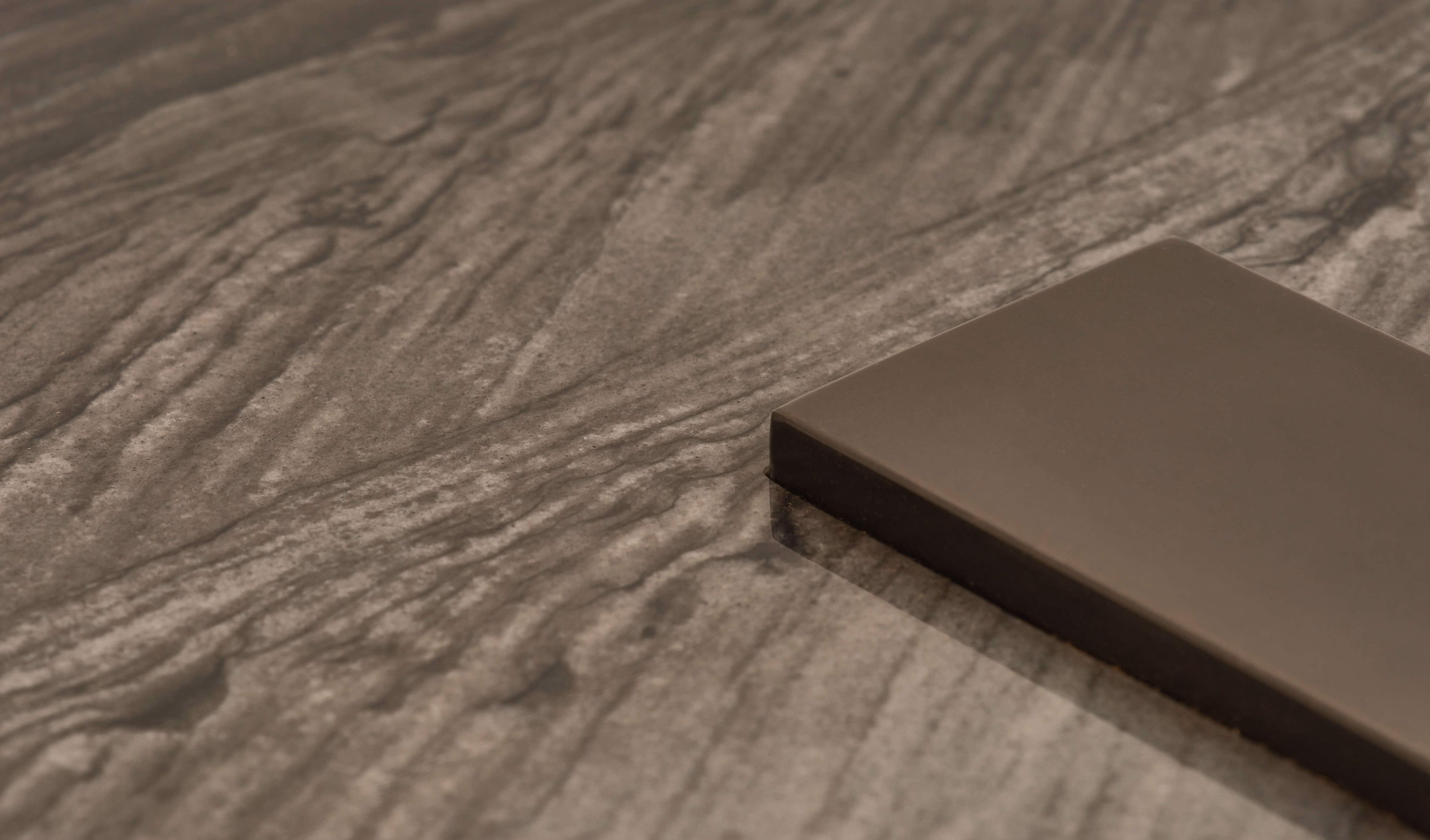 Close-up of a dark walnut wood slab textured with visible grain patterns and a smooth, rectangular dark brown material sample resting on top.