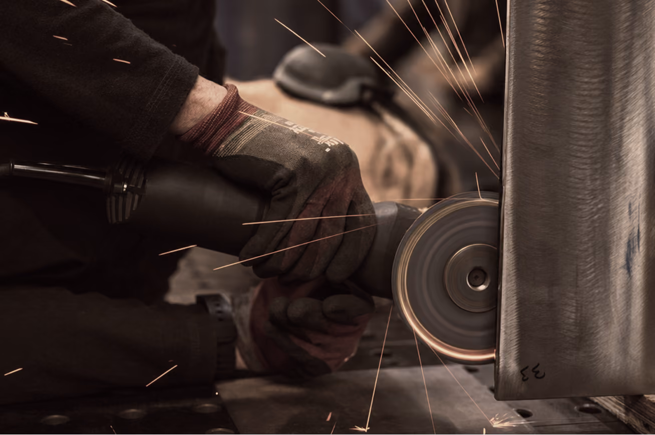 Person wearing gloves using a handheld grinder to cut or polish a metal surface with sparks flying.