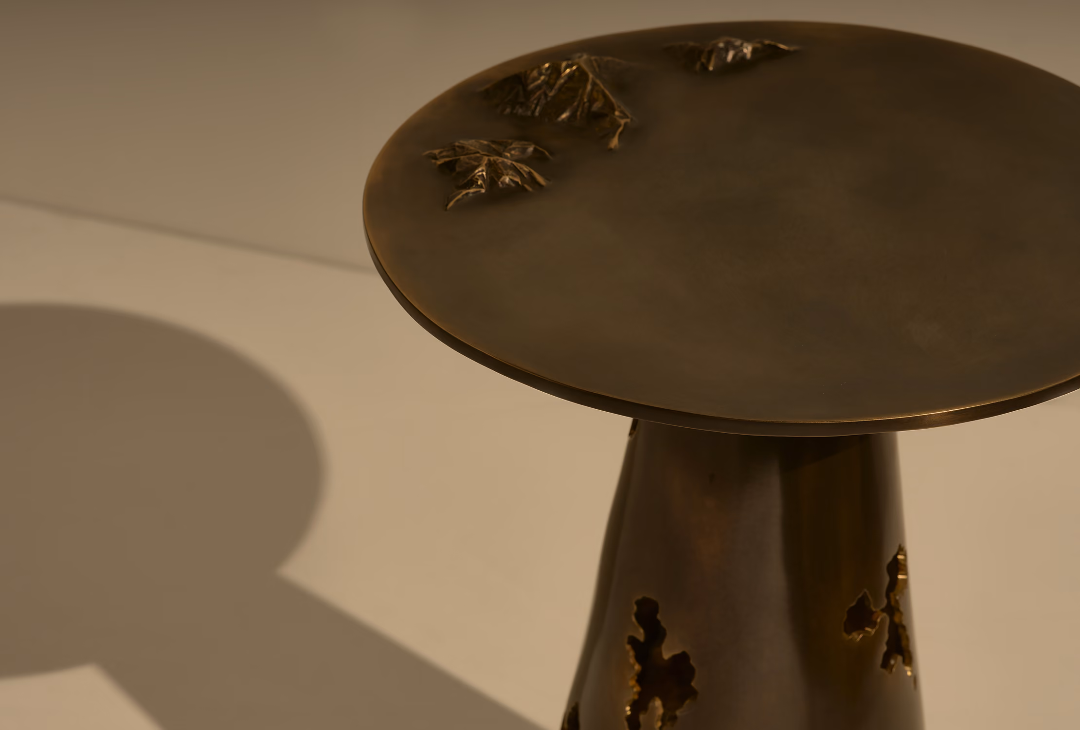 Close-up of a bronze or dark metal side table with a round top and an artistic design featuring textured, irregularly-shaped cutouts and raised leaf-like details on the surface, casting a shadow on a light background.