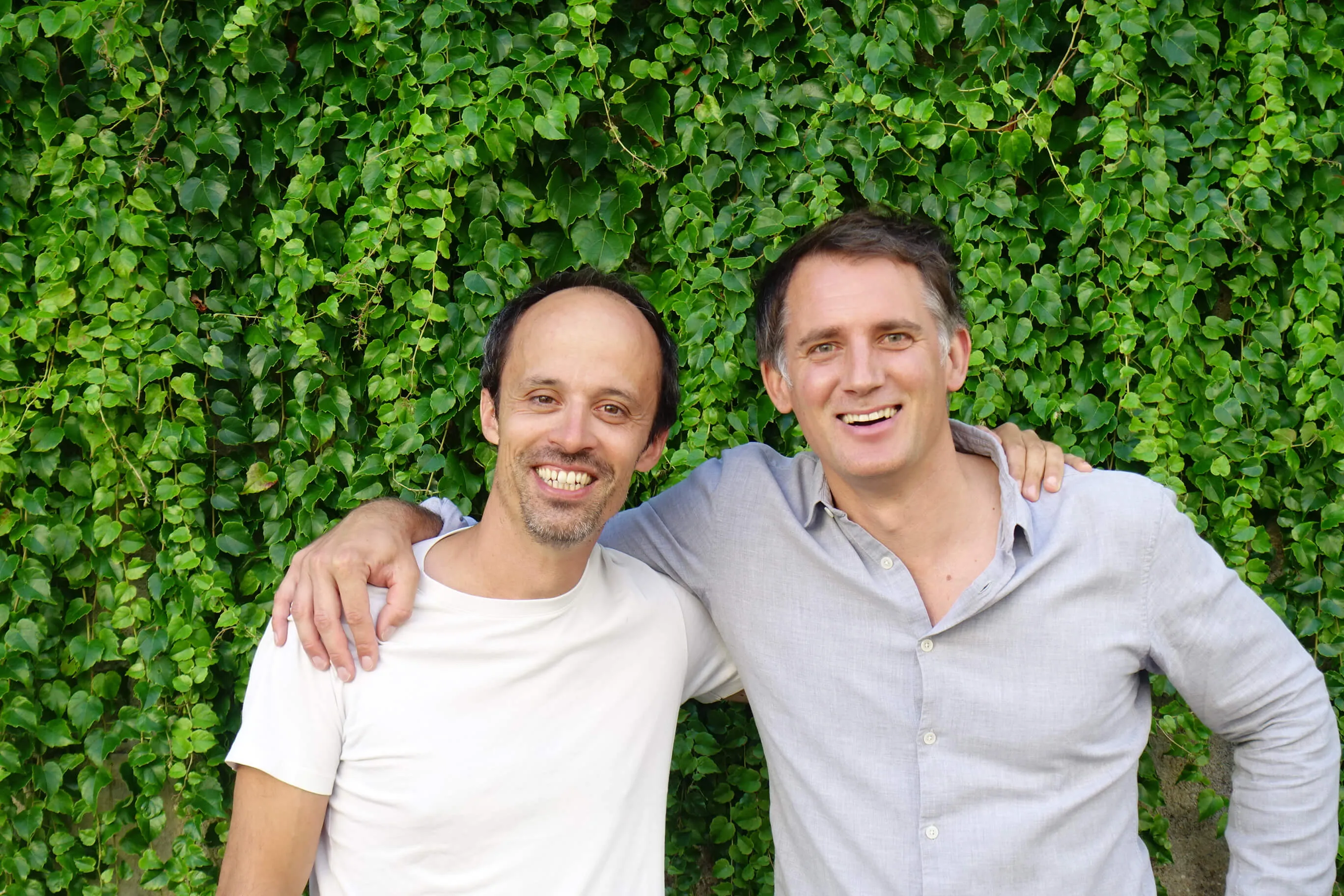 Deux hommes souriants, bras sur les épaules, devant un mur couvert de feuilles vertes.