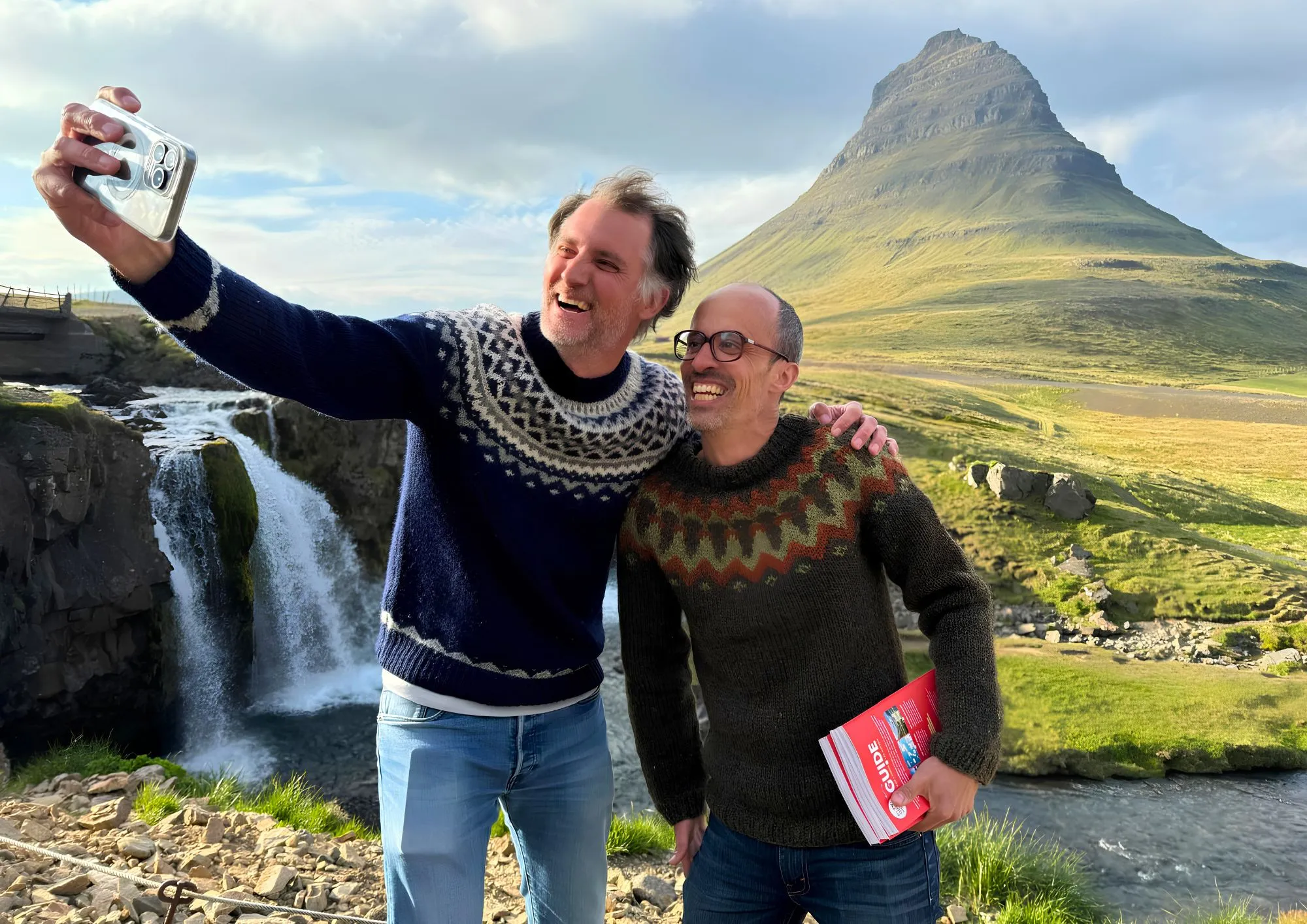 Deux hommes souriants prennent un selfie devant une cascade et une montagne en Islande.