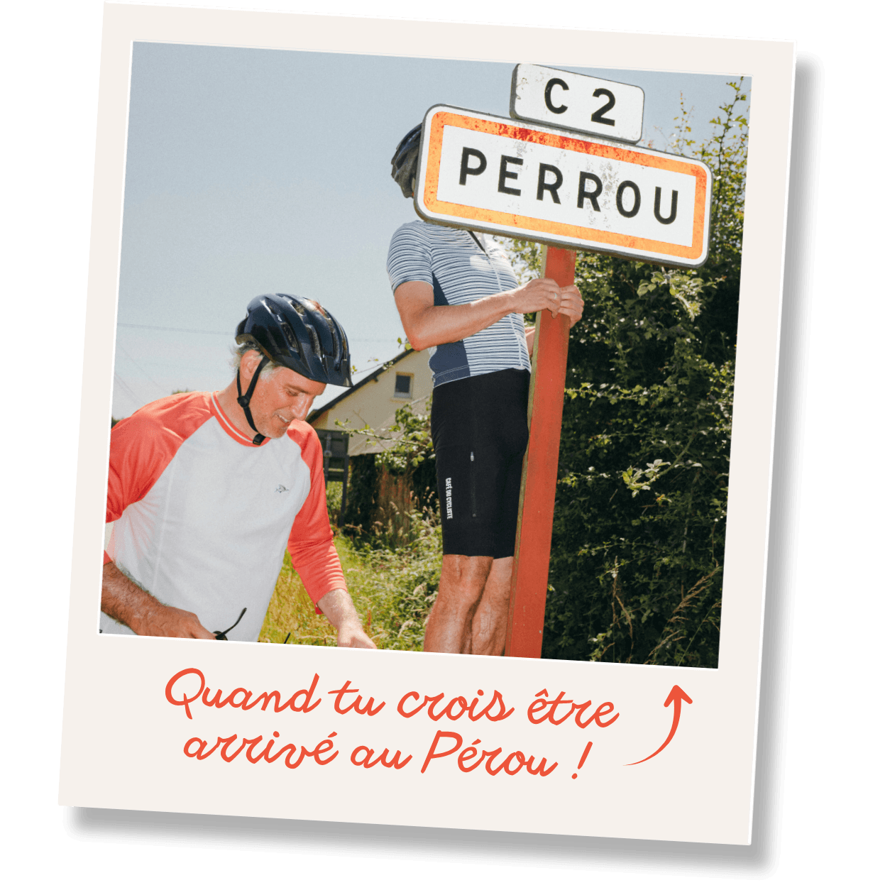 Deux cyclistes posent près d'un panneau indiquant Perrou, sur fond de ciel clair et végétation.