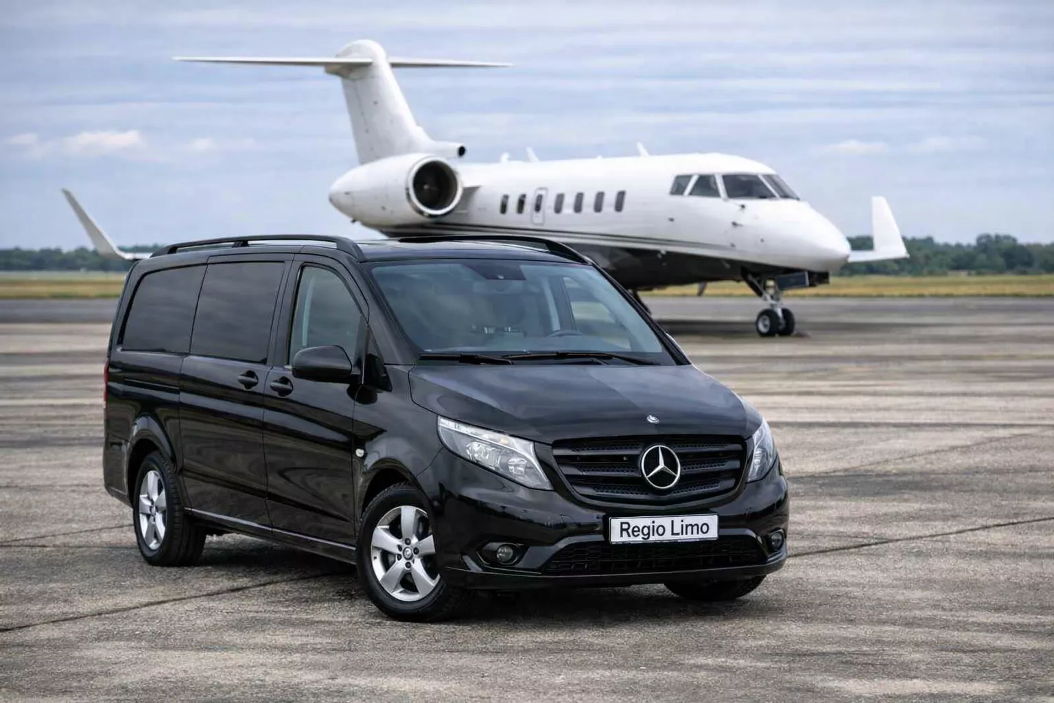 Black Mercedes van with Regio Limo license plate parked on an airport tarmac with a white private jet in the background.