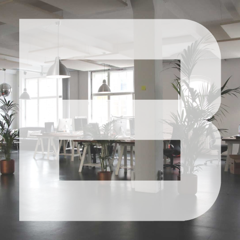 Modern office space with desks, computers, hanging lights, and large indoor plants.