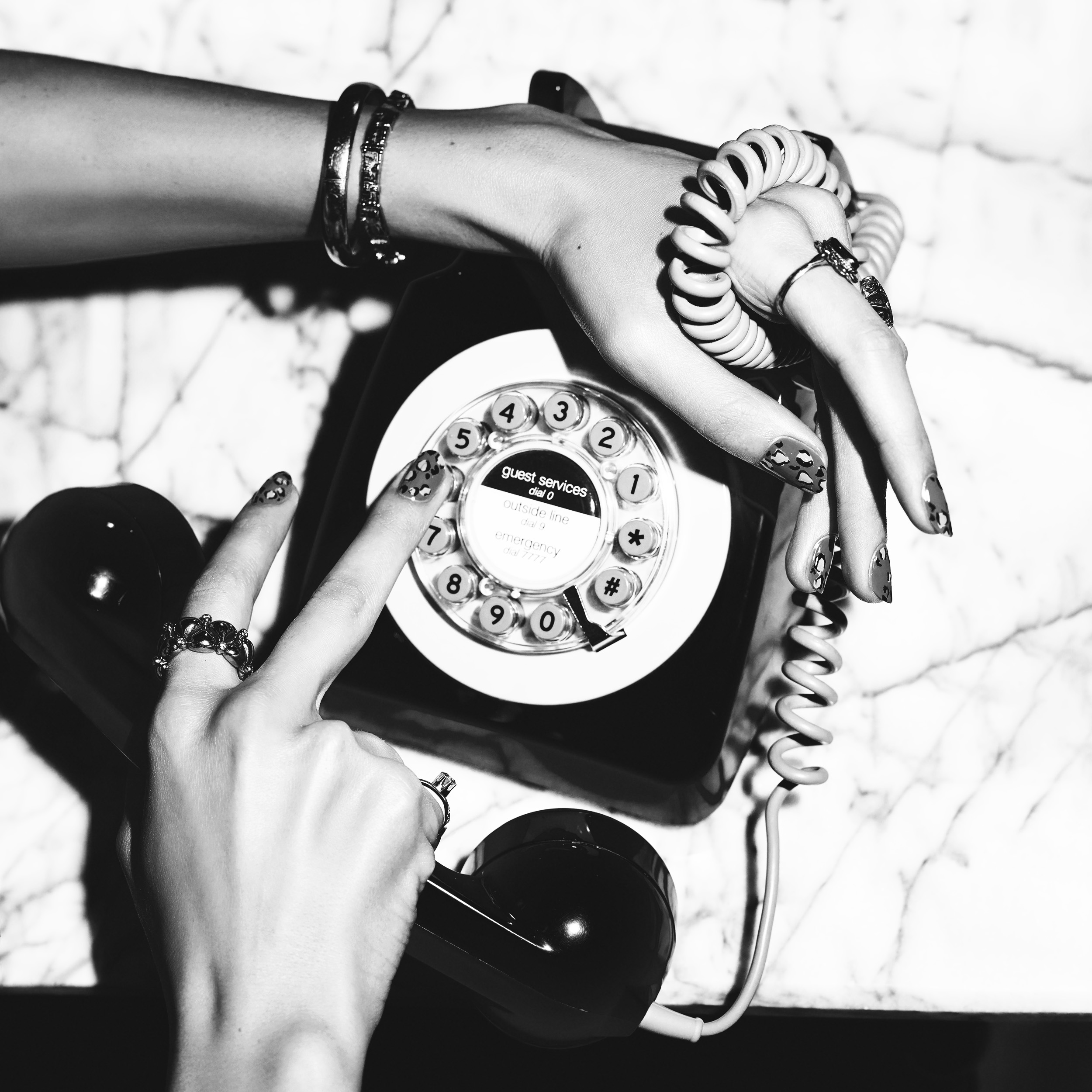 Two hands dialing a black rotary phone on a marble surface, with rings and bracelets worn.