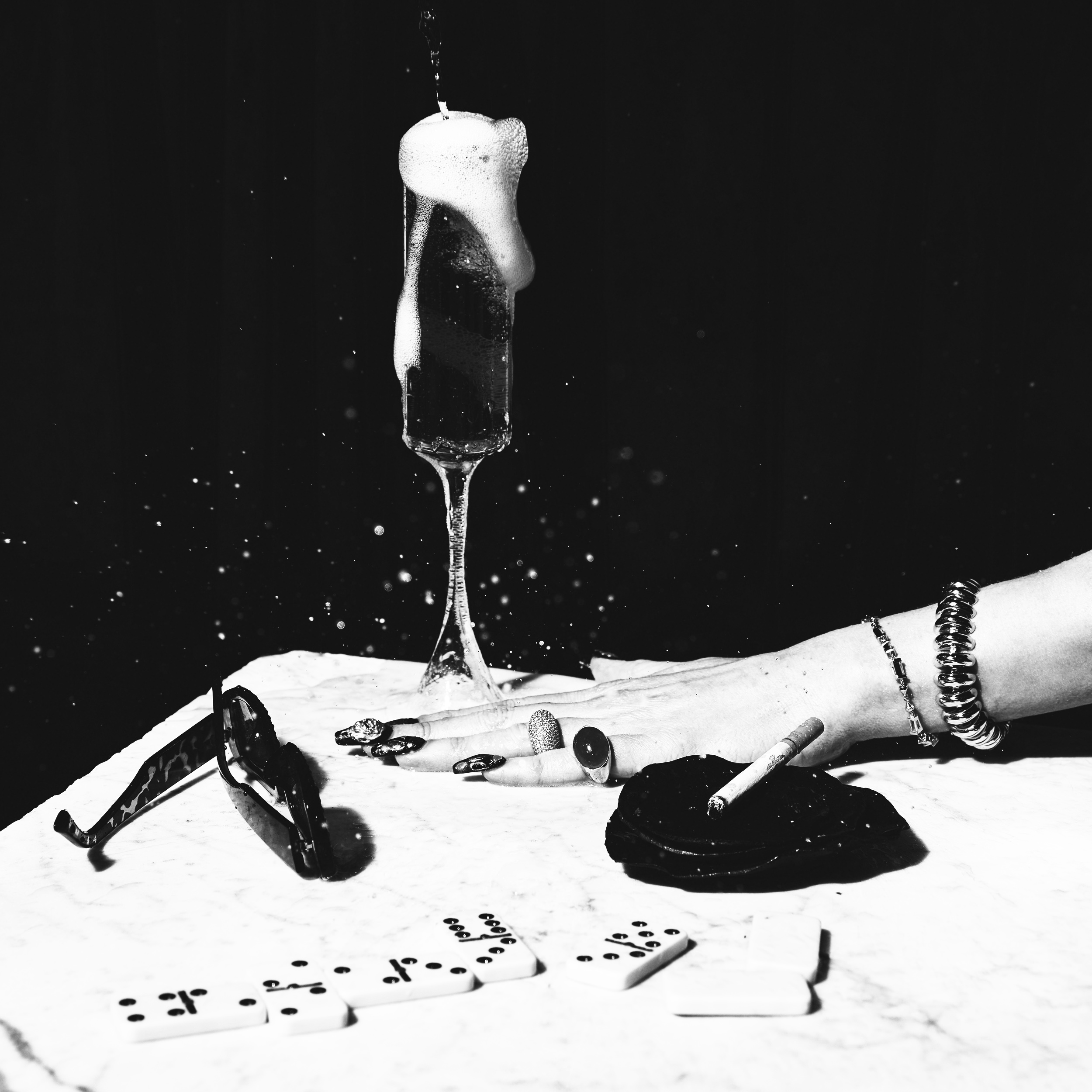 Hand with rings and bracelets next to sunglasses, dominoes, a cigarette, and a champagne glass overflowing with foam.