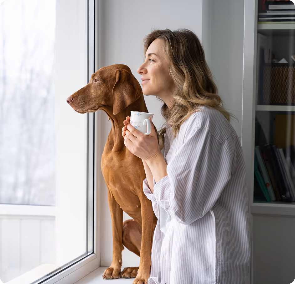 A woman in a light striped shirt holding a cup, standing beside a brown dog as they both look out a window.