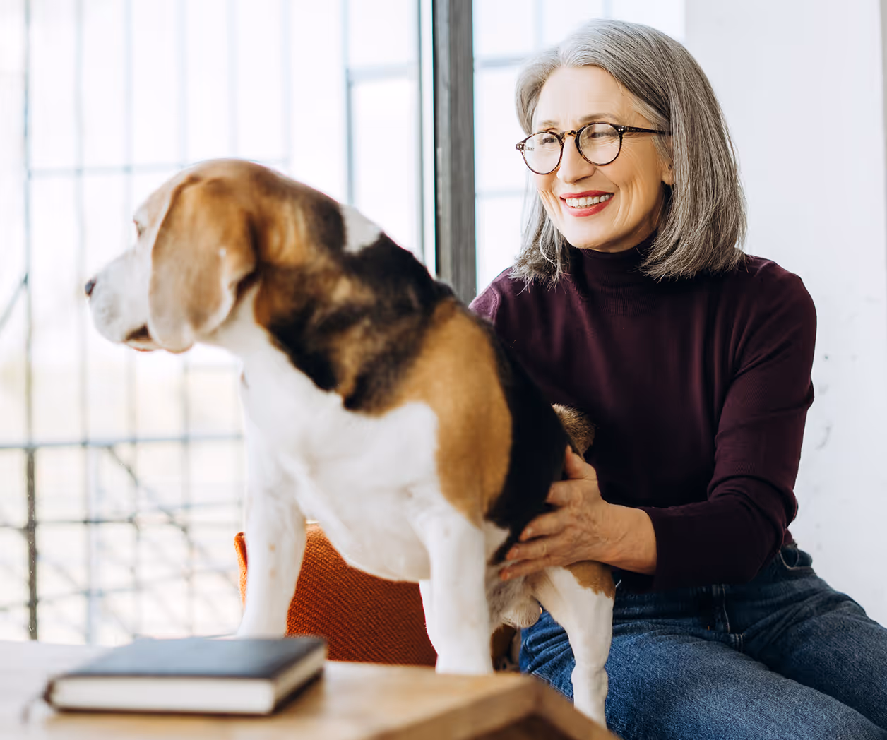 Smiling woman with gray hair and glasses sitting indoors petting a beagle dog beside a table with a closed book.