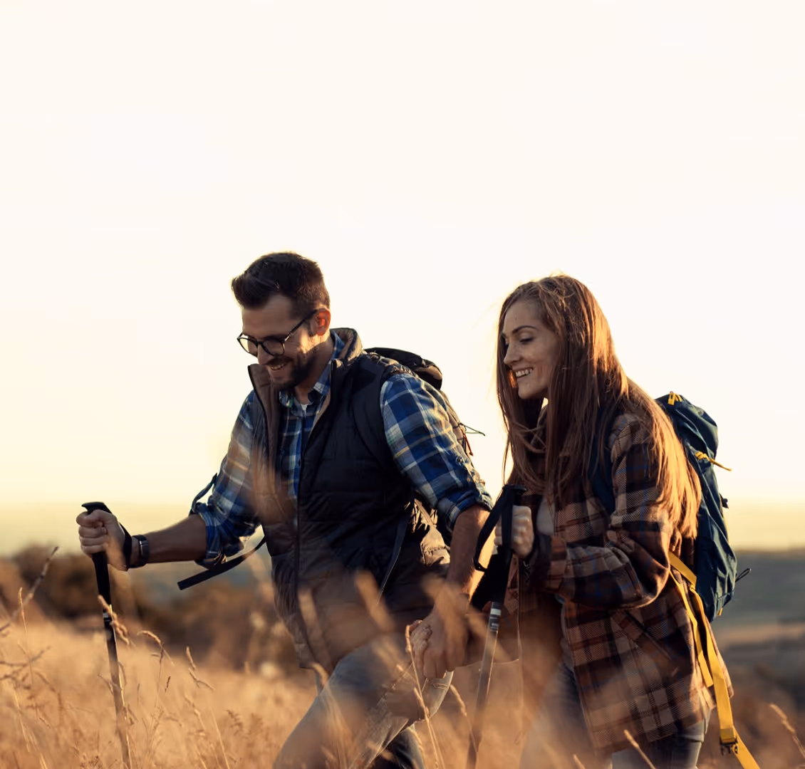 Smiling man and woman hiking outdoors in a grassy field at sunset, holding hands and using trekking poles.