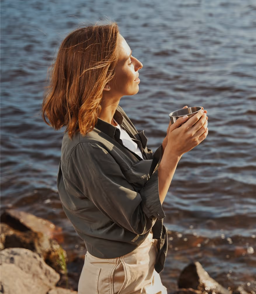 Woman with closed eyes holding a metal cup, standing by a rocky shoreline with calm water.