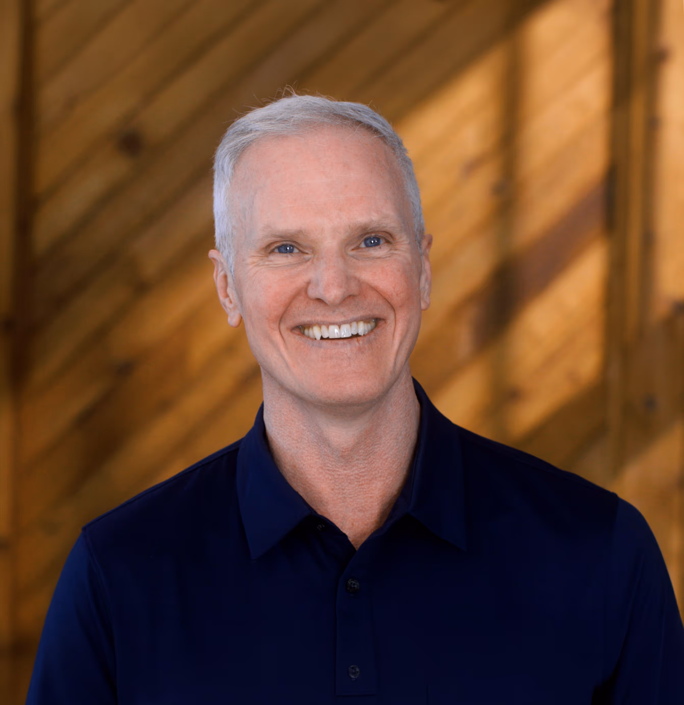 Smiling middle-aged man with short gray hair wearing a navy blazer and white shirt against a blurred sunset background.