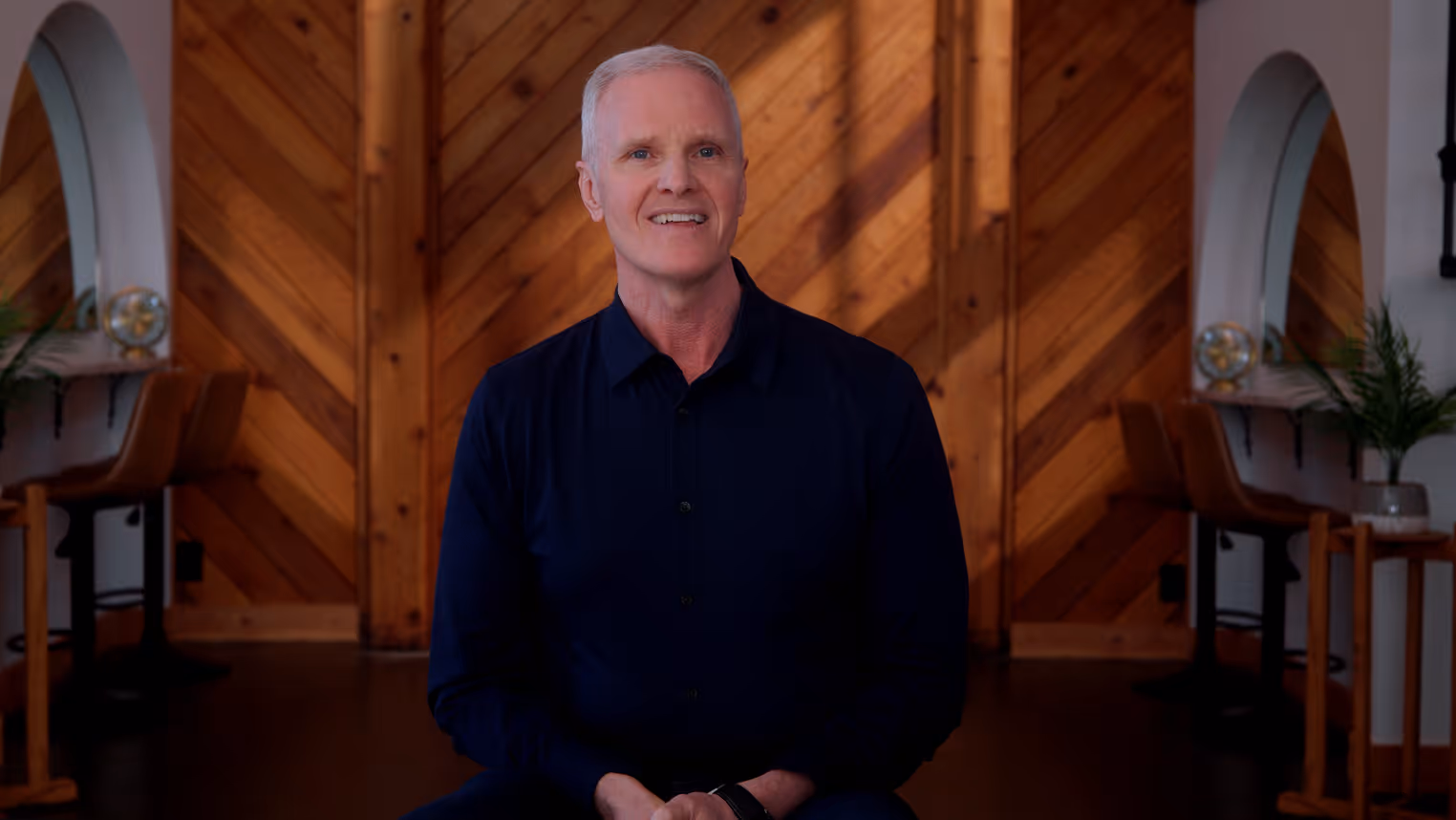 Smiling middle-aged man with short gray hair wearing a dark shirt sitting in front of a wood-paneled wall in a warmly lit room.