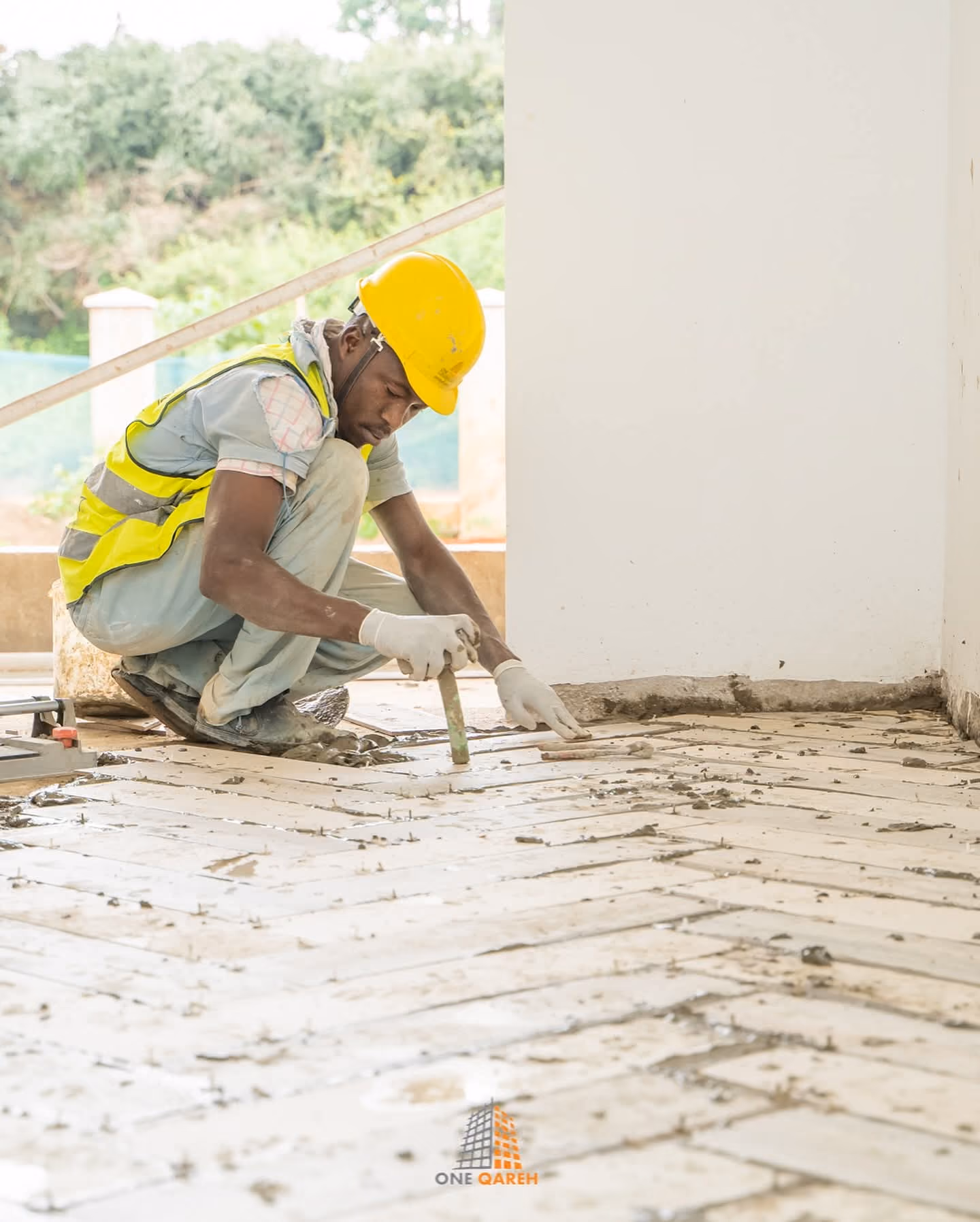 Construction worker in yellow helmet installing floor tiles indoors.