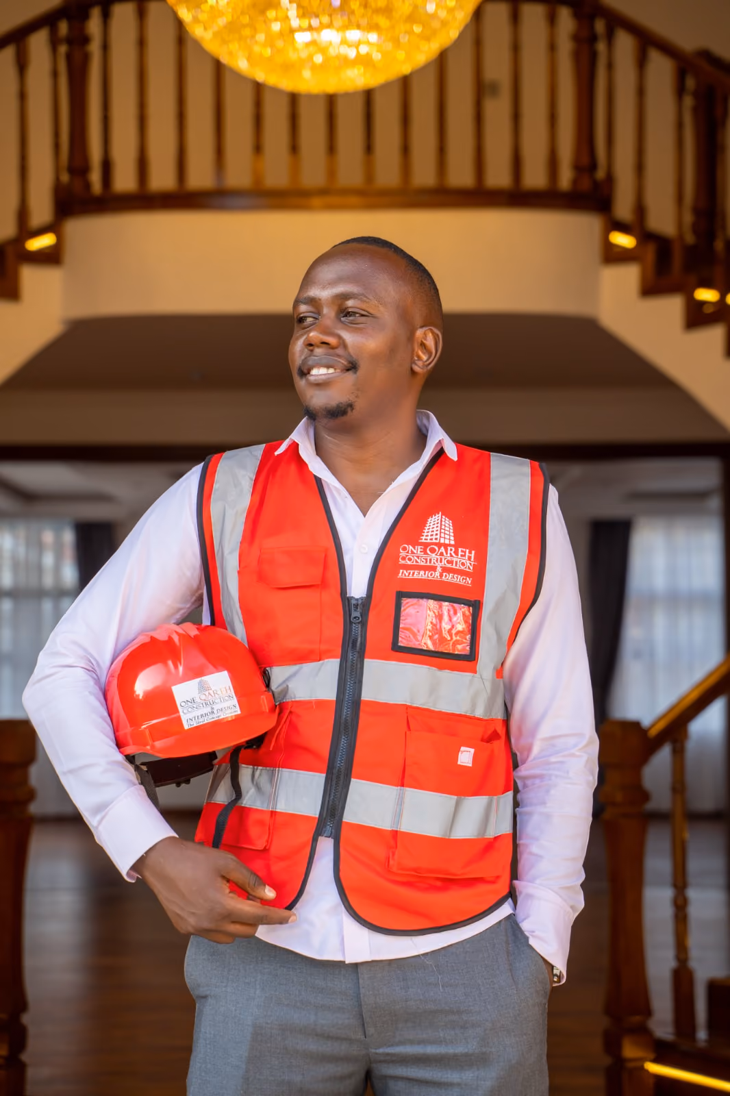 Man in a red construction vest holding a red hard hat inside a building with wooden staircase