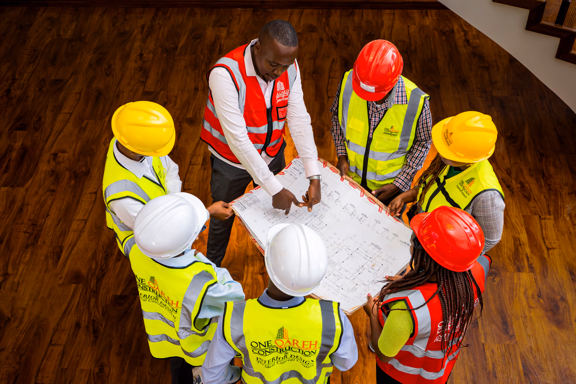 Group of construction workers in helmets and vests reviewing blueprints together indoors.
