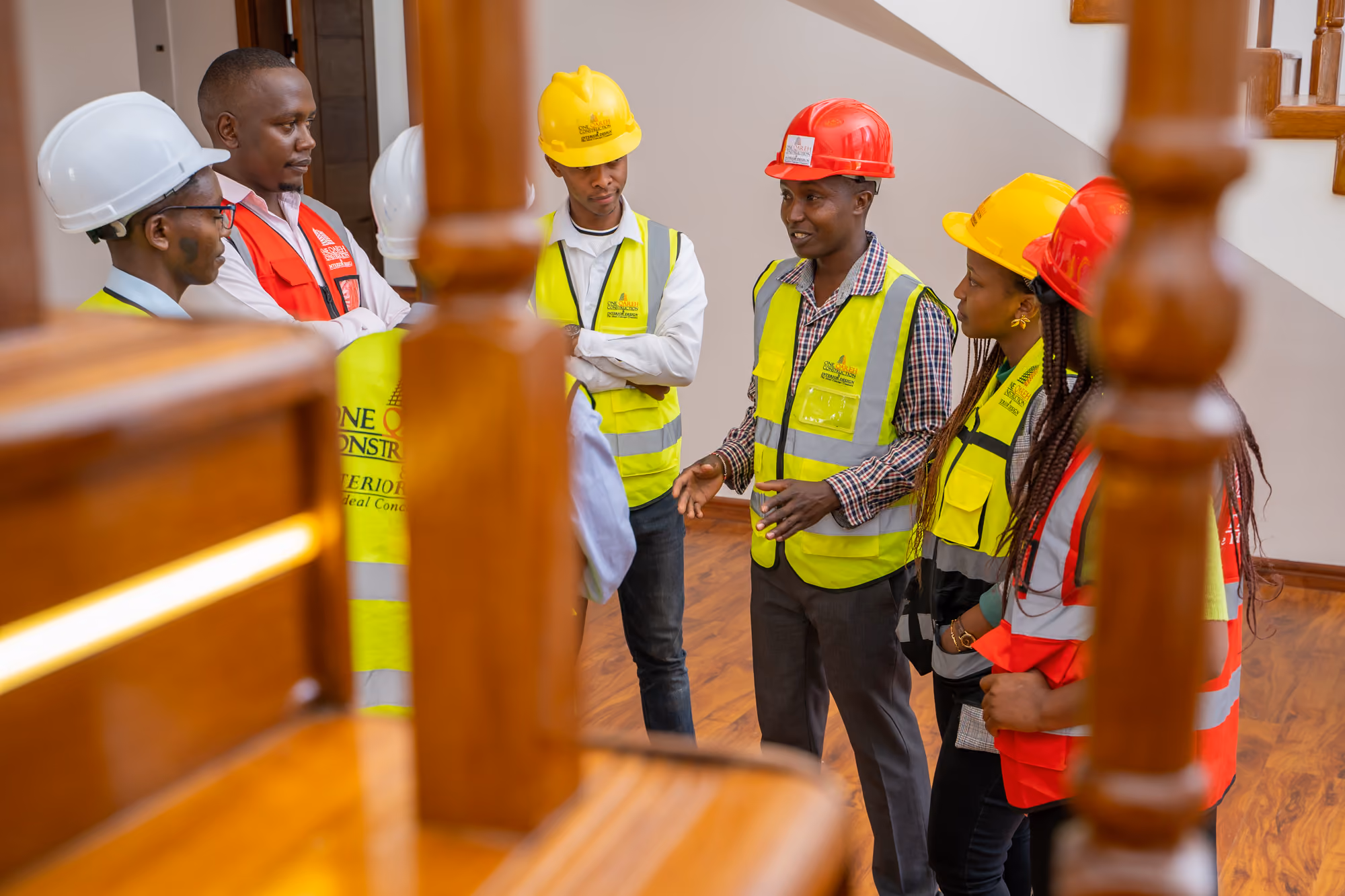 Group of construction workers in safety vests and helmets discussing indoors near a wooden staircase.