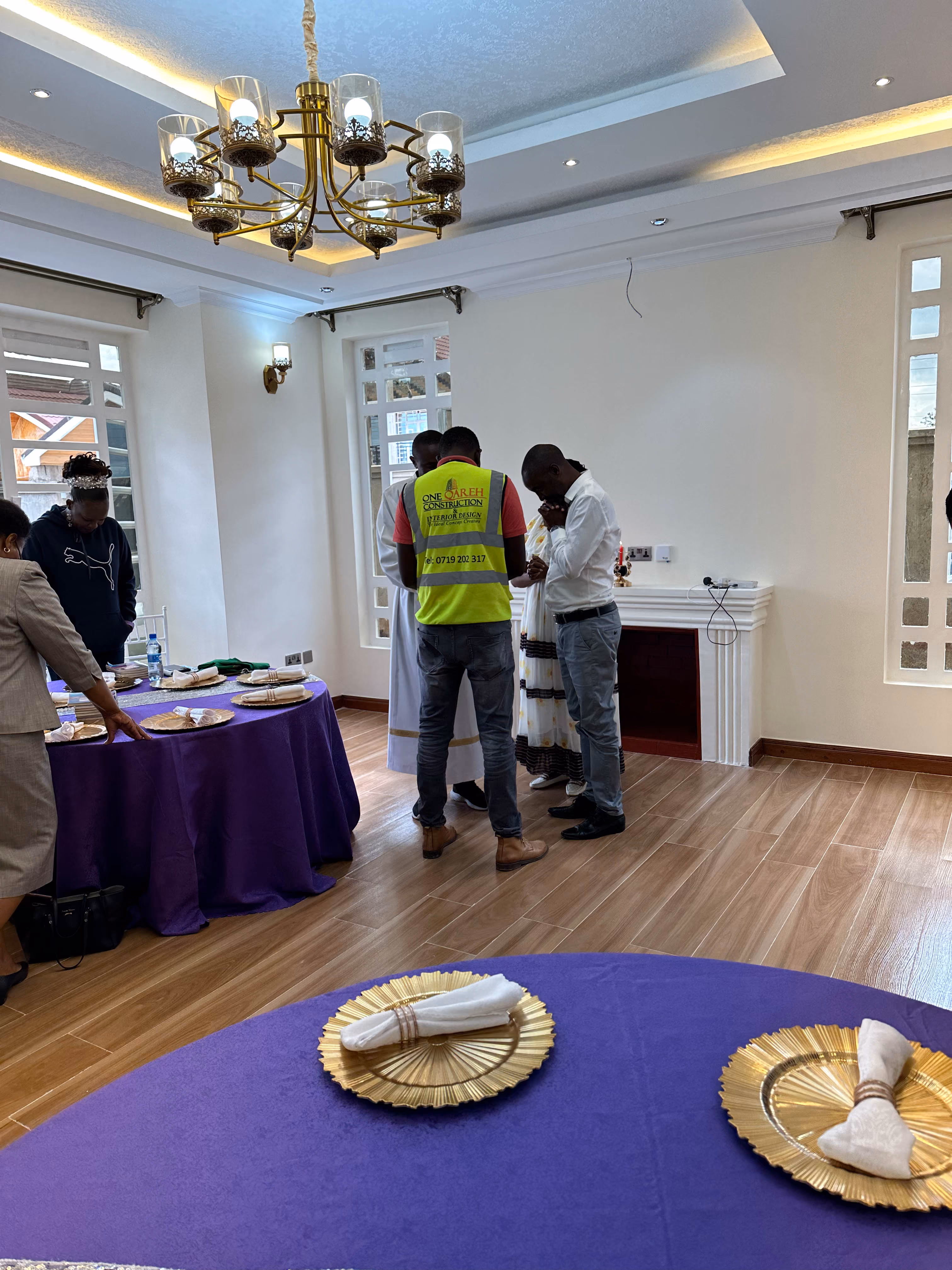 People standing and praying in a decorated room with purple tables and gold plates.