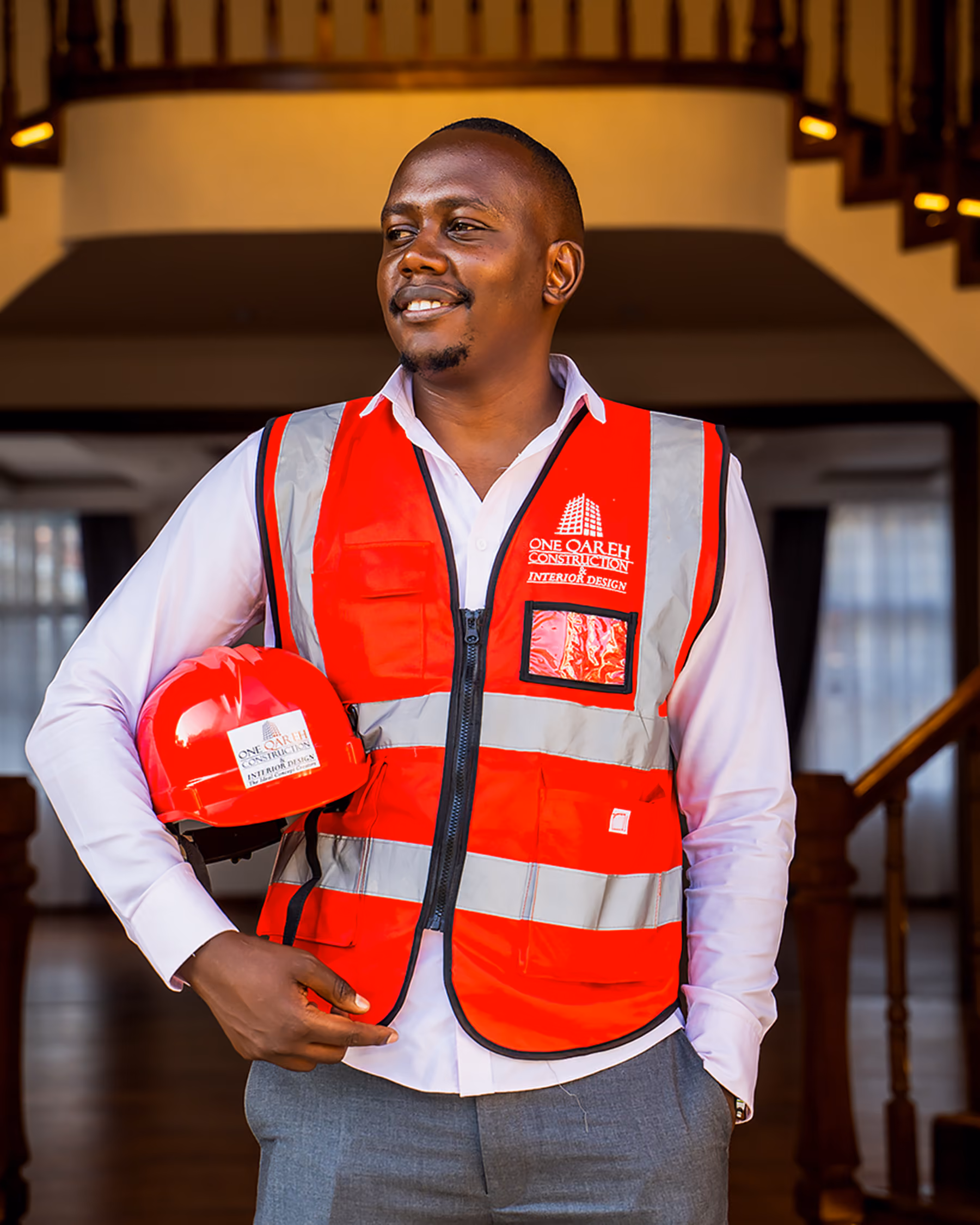 Smiling man in a red construction safety vest holding a red hard hat indoors by stairs.
