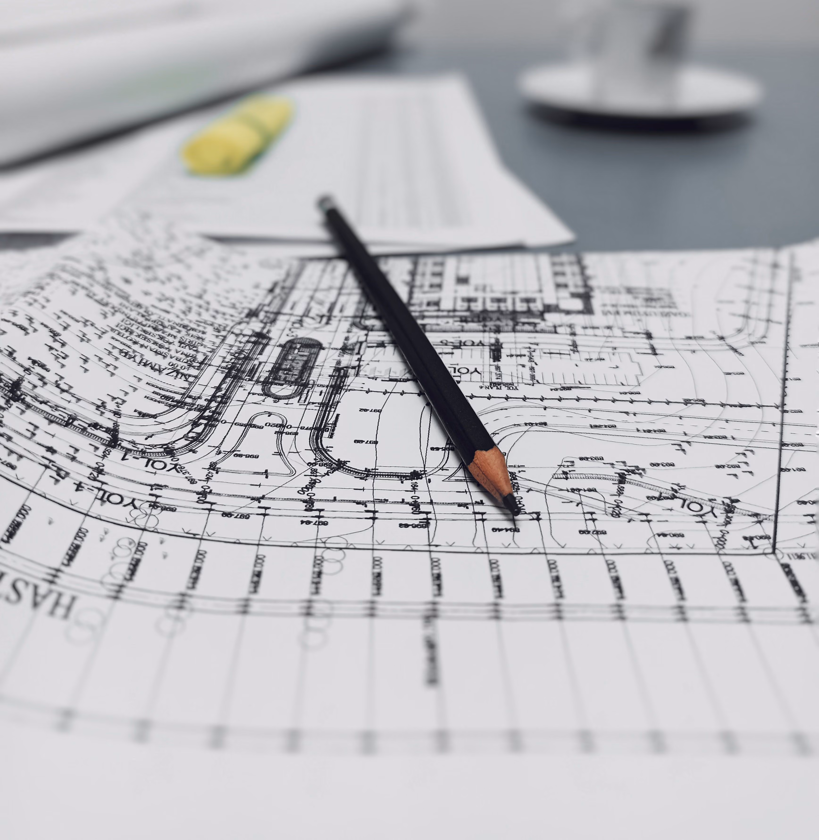 Black pencil resting on architectural blueprints with blurred papers and a cup in the background.