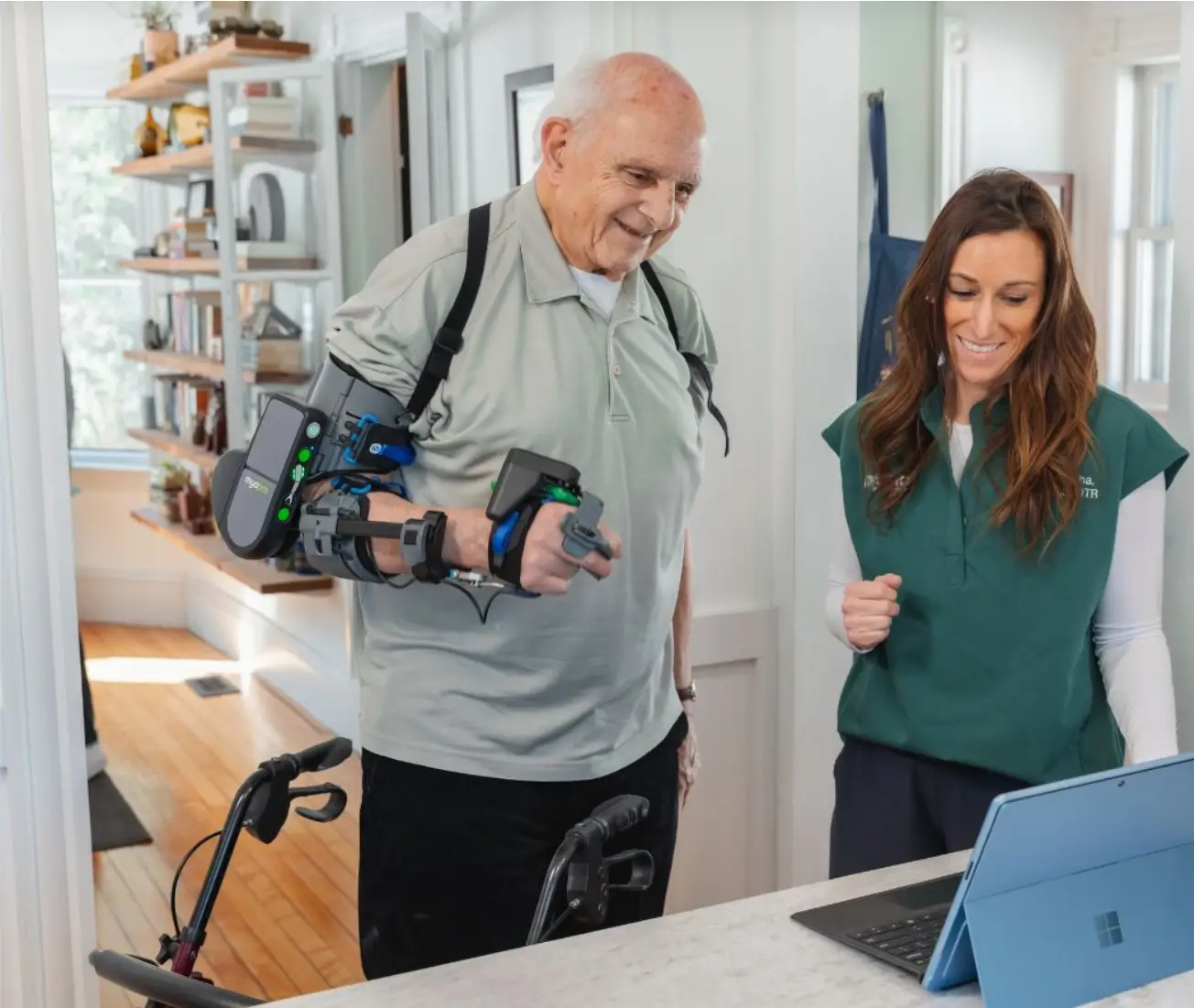 Senior man wearing a robotic arm exoskeleton device being assisted by a smiling woman with a laptop.