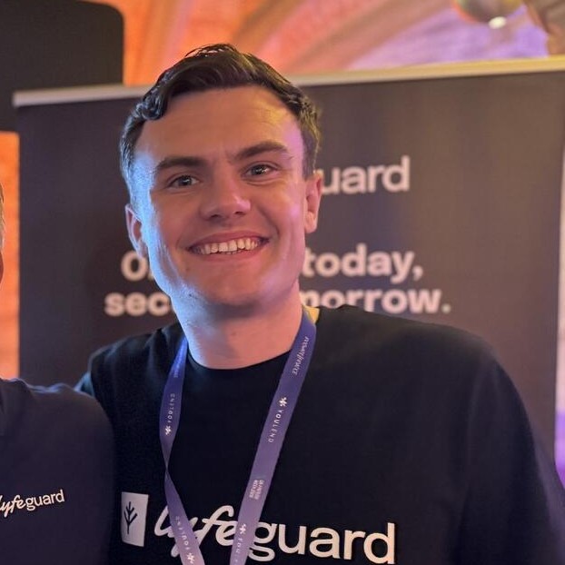 Smiling man wearing a lifeguard shirt and a blue lanyard at an event with blurred background.