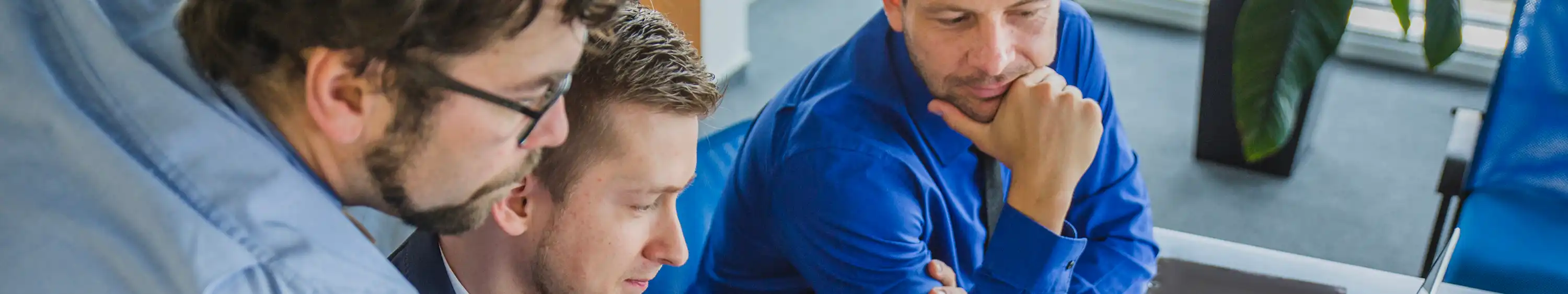Three men in office attire closely collaborating over a desk with laptops and documents.