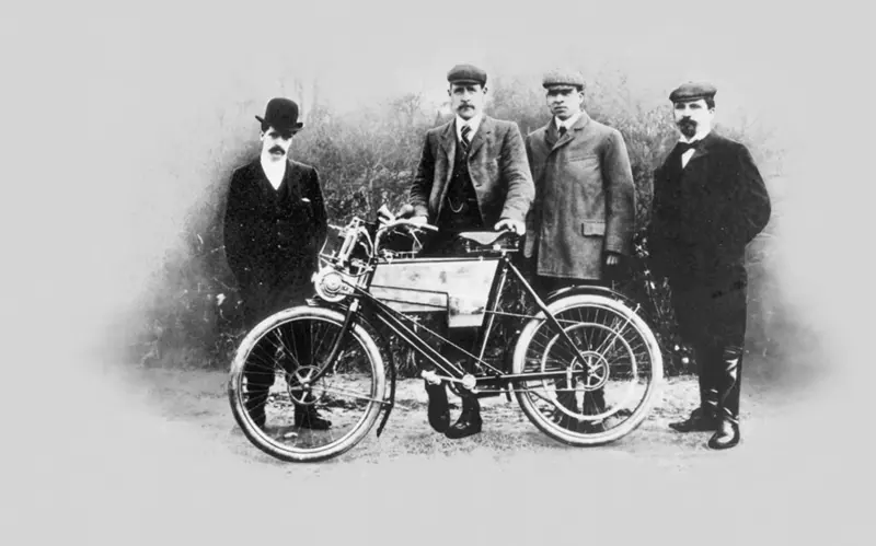 Čtyři muži v kloboucích pózují se starým motorovým kolem z počátku 20. století.