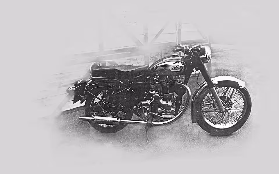 Černobílý snímek klasické motorky Royal Enfield stojící na stojanu vedle dřevěné konstrukce.