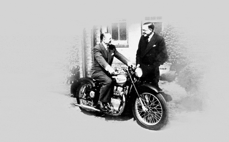 Černobílá historická fotografie dvou mužů v oblecích; jeden sedí na starém motocyklu a druhý stojí vedle něj.