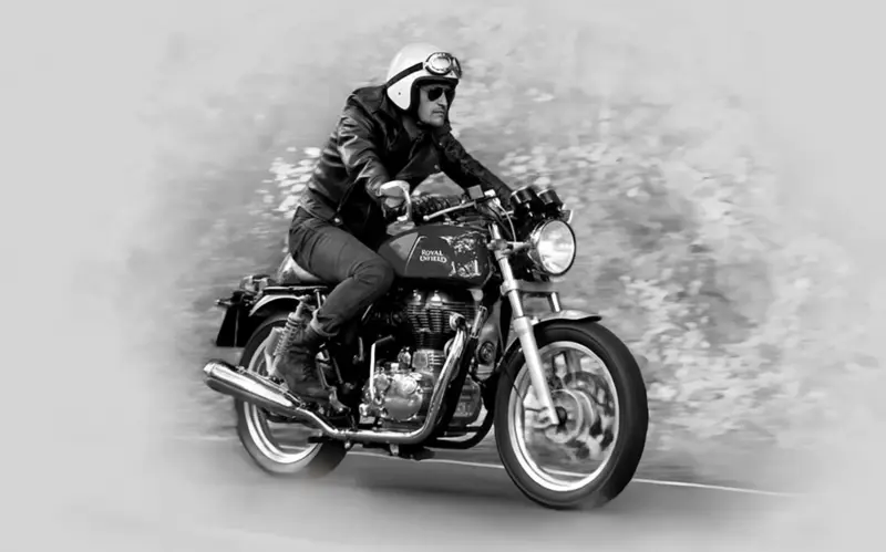 Muž v kožené bundě a přilbě jede na černé motorce Royal Enfield po silnici.