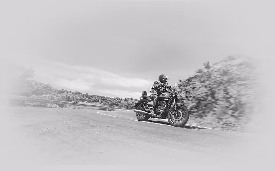 Jezdec na motocyklu jedoucím po venkovské silnici s krajinou a stromy v pozadí.
