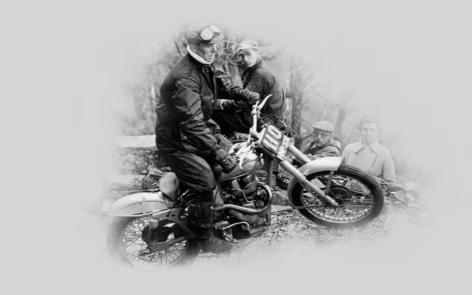 Černobílá fotografie jezdce na terénním motocyklu během závodu, v pozadí dva sedící muži a jeden stojící.