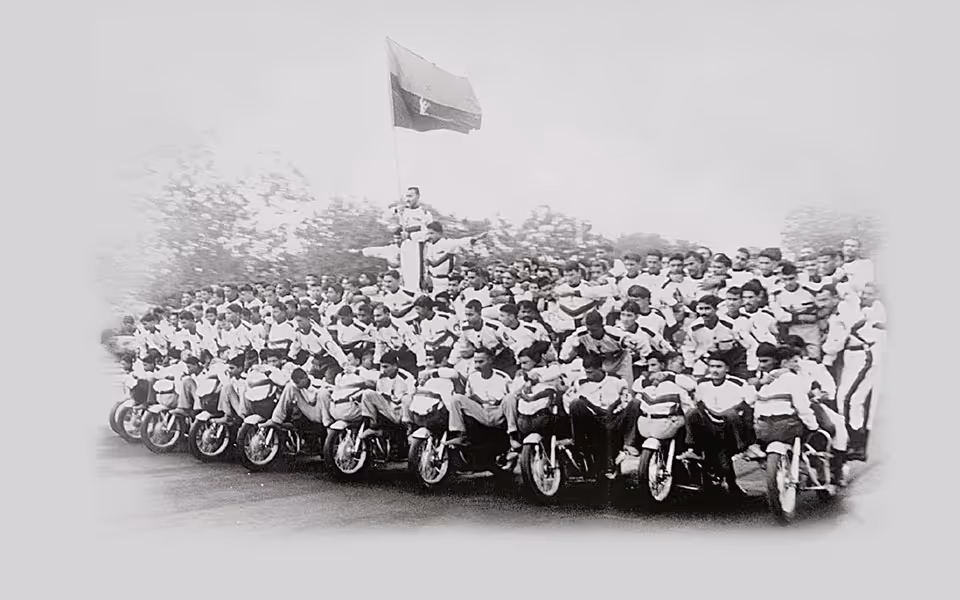 Velká skupina mužů sedících a stojících na motocyklech v uspořádané formaci s vlajkou na pozadí.